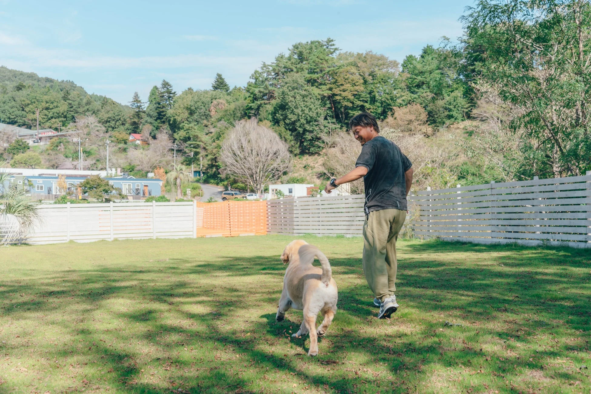 晴れた日に、男性が広い芝生のドッグランでゴールデンレトリバーと一緒に散歩したり遊んだりしている様子