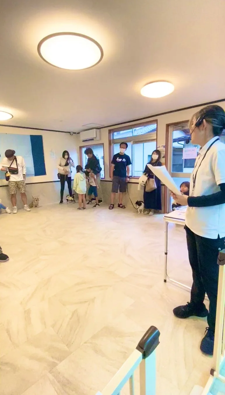 室内でペットを連れた家族連れが集まっている様子です。ヘッドセットをつけた女性が何か説明しているようで、犬を伴った人々が耳を傾けています。