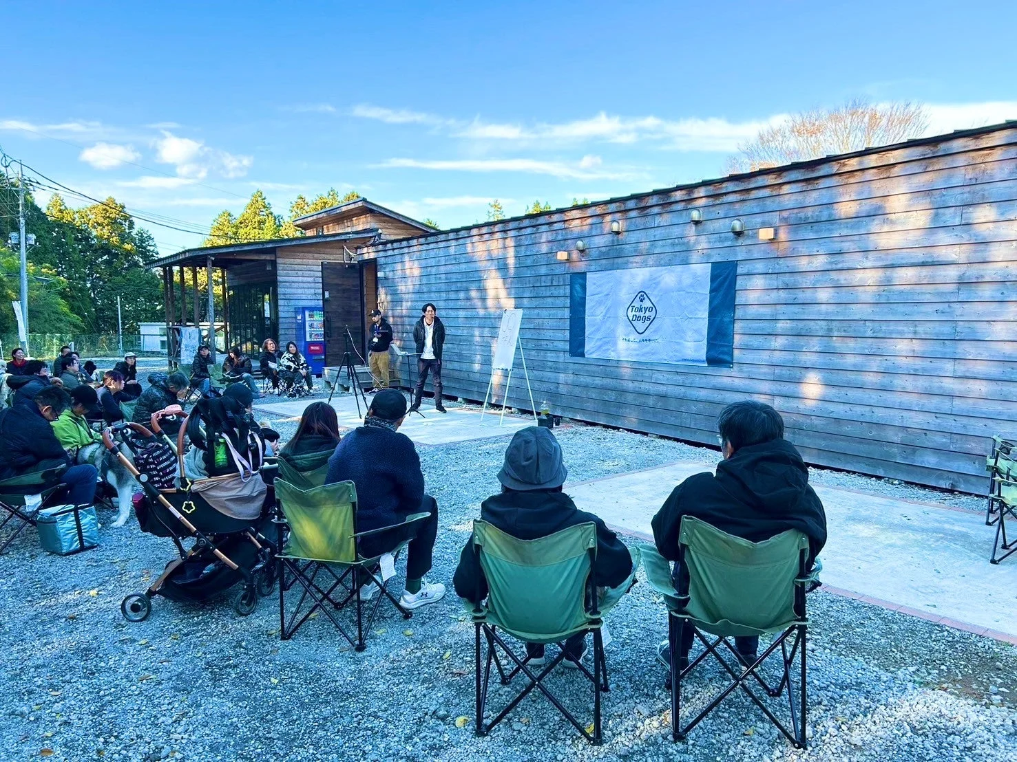 青空の下、屋外で人々がキャンプチェアに座り、プレゼンターの話を聞いている様子です。犬と暮らす幸せをテーマにした「Tokyo Dogs」のイベントで、和やかなコミュニティの集まりに見えます。